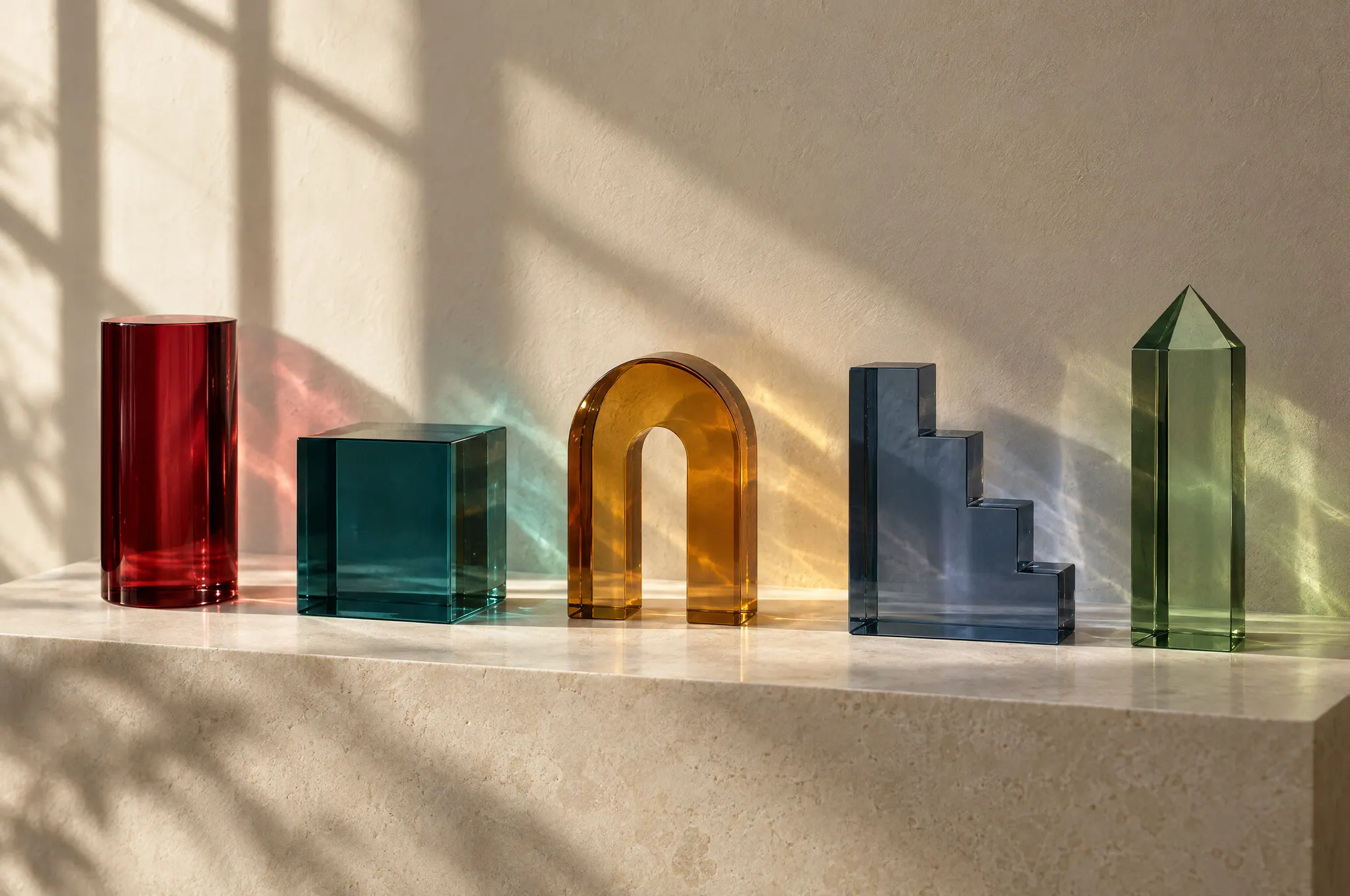 Illustrative still life of five glass forms in five colors, side by side on a stone ledge.